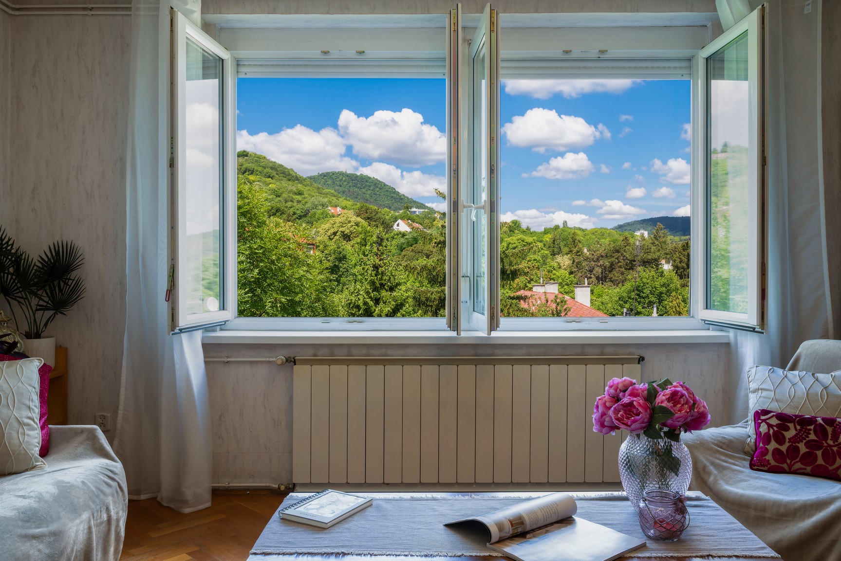Nyitott ablakon át látható nyári panoráma, amely a fényt, a levegősséget és az otthonérzetet mutatja meg.
